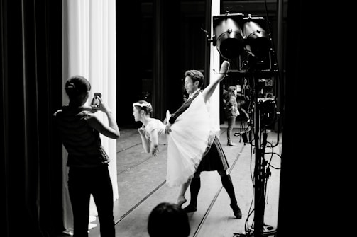 A dance performance taking place backstage, where a male dancer holds a female dancer in a lifted pose. A person in the foreground captures the moment with a smartphone. Stage lighting and equipment are visible in the scene.