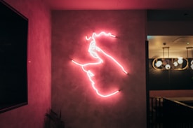 Neon pink outlines of a face and hand are mounted on a wall, creating a striking light installation with a contemporary aesthetic. The setting suggests an interior space with modern lighting fixtures.