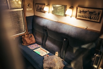 A cozy train compartment with blue cushioned seats, softly lit by two wall lamps. There are framed pictures on the wooden walls and a window reflecting the inside lights. Various items like a handbag, a sweater, and a few magazines or newspapers are placed on the seat.