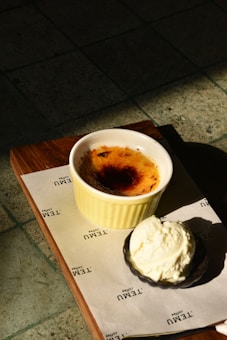 A wooden tray holds a dessert setup, featuring a ramekin of cr&egrave;me br&ucirc;l&eacute;e with a caramelized top and a side of vanilla ice cream in a small black bowl. The tray sits on a piece of paper with 'TEMU Coffee' printed on it, placed on a tiled surface, partially illuminated by sunlight.