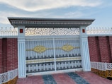 An intricate wrought iron gate adorned with golden and black accents. The gate is flanked by white columns with vertical lines and topped with a decorative metal pattern. The surrounding wall is made of red bricks with white and gold decorative fencing on top. The ground is tiled with a mixture of red and gray tiles, and the sky above is mostly clear with some light clouds.