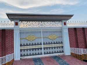 An intricate wrought iron gate adorned with golden and black accents. The gate is flanked by white columns with vertical lines and topped with a decorative metal pattern. The surrounding wall is made of red bricks with white and gold decorative fencing on top. The ground is tiled with a mixture of red and gray tiles, and the sky above is mostly clear with some light clouds.