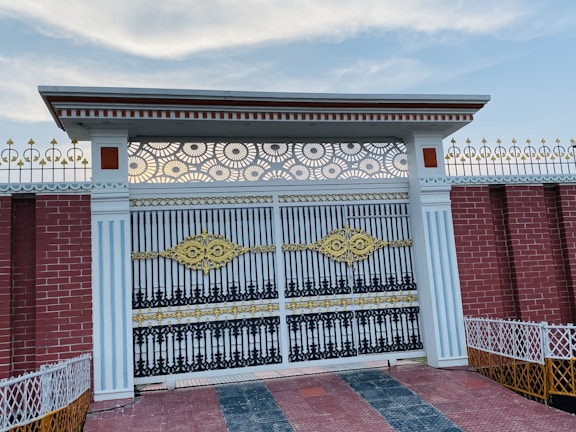 An intricate wrought iron gate adorned with golden and black accents. The gate is flanked by white columns with vertical lines and topped with a decorative metal pattern. The surrounding wall is made of red bricks with white and gold decorative fencing on top. The ground is tiled with a mixture of red and gray tiles, and the sky above is mostly clear with some light clouds.
