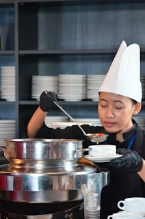 A chef wearing a white hat and black gloves is pouring soup from a large stainless steel pot into a white bowl. Shelves filled with stacks of white plates can be seen in the background.