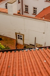 An aerial view of a section of red-tiled roofs with scattered debris, including cans and small objects. The scene is surrounded by white building walls with several windows and a small door. The overall setting gives an impression of wear and neglect.