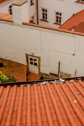 An aerial view of a section of red-tiled roofs with scattered debris, including cans and small objects. The scene is surrounded by white building walls with several windows and a small door. The overall setting gives an impression of wear and neglect.