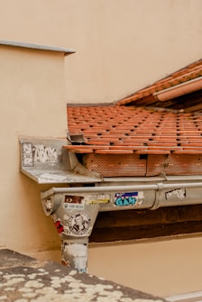 A section of a rooftop with reddish-brown tiles and a metal gutter system. The gutter is covered with multiple colorful stickers and graffiti, contrasting with the weathered, off-white wall and roofing tiles.