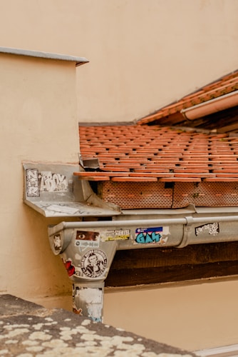 A section of a rooftop with reddish-brown tiles and a metal gutter system. The gutter is covered with multiple colorful stickers and graffiti, contrasting with the weathered, off-white wall and roofing tiles.