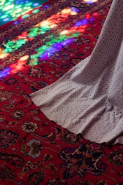 A beautifully patterned red carpet with intricate designs is illuminated by colorful light reflections. Part of a draped fabric flows across the carpet, adding to the rich texture and color.