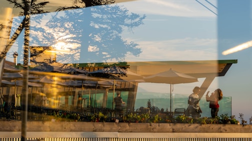 A reflection displays two people interacting on a terrace with outdoor umbrellas and seating. The setting is enhanced by the reflections of trees and a sunset, creating a layered view with natural elements blending into the café environment.