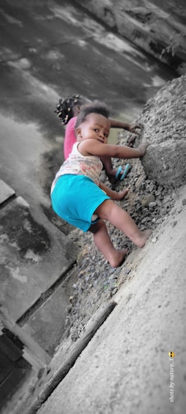 A young child is climbing a rocky slope with determination, dressed in a colorful outfit. Another child is partially visible in the background, focused on their activity. The scene appears to be outdoors against a textured wall.