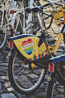 A brightly colored bicycle with a yellow panel featuring a heart with rainbow colors and the text 'Wir bewegen alle.' is parked on a cobblestone path among other bicycles. The surrounding bicycles are mostly white, and the ground is covered with cobblestone and some moss.