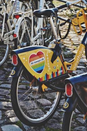 A brightly colored bicycle with a yellow panel featuring a heart with rainbow colors and the text 'Wir bewegen alle.' is parked on a cobblestone path among other bicycles. The surrounding bicycles are mostly white, and the ground is covered with cobblestone and some moss.