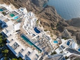 An aerial view of a luxurious coastal resort with multiple swimming pools. The resort features white buildings with terraces and sun loungers, nestled into rocky cliffs overlooking a blue ocean. The landscape is a blend of architectural elegance and natural beauty.