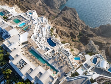 An aerial view of a luxurious coastal resort with multiple swimming pools. The resort features white buildings with terraces and sun loungers, nestled into rocky cliffs overlooking a blue ocean. The landscape is a blend of architectural elegance and natural beauty.