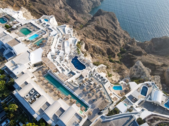 An aerial view of a luxurious coastal resort with multiple swimming pools. The resort features white buildings with terraces and sun loungers, nestled into rocky cliffs overlooking a blue ocean. The landscape is a blend of architectural elegance and natural beauty.