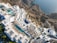 An aerial view of a luxurious coastal resort with multiple swimming pools. The resort features white buildings with terraces and sun loungers, nestled into rocky cliffs overlooking a blue ocean. The landscape is a blend of architectural elegance and natural beauty.