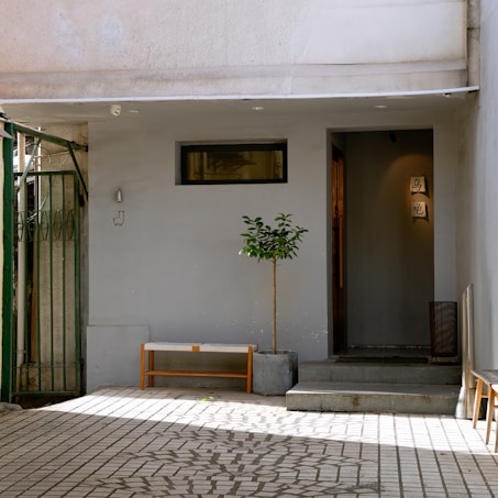 A minimalist exterior with a small potted tree in front of a modern building entrance. The façade is simple, with a rectangular window and a small light fixture. A wooden bench is placed along the wall, and there are two signs with characters hanging near the doorway. The area is paved with patterned tiles, casting shadows from nearby structures.