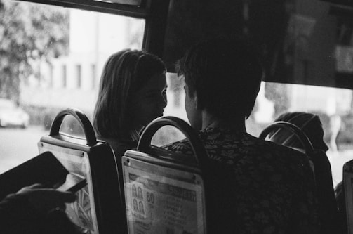 A black and white photo capturing two people seated closely together on a bus or train, with one turning slightly to face the other. The image has a candid, intimate feel, suggesting a private conversation or moment. The surroundings include seats and windows typical of public transportation.