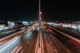 A highway at night with light trails from fast-moving vehicles on both sides. The road is separated by a barrier with a light pole in the center. Buildings and shops line the roadside, illuminated by various lights and signs, creating a vibrant urban atmosphere.