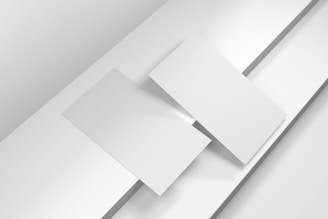 Two overlapping blank white sheets are placed atop a layered background of lighter white and grey surfaces, creating a minimalist and geometric composition.