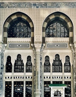 An intricate and ornate architectural facade featuring large arched windows with decorative patterns. The structure is adorned with geometric and floral motifs, displaying detailed craftsmanship in stone and glass. The design combines traditional and artistic elements, emphasized by columns and arches, creating a sense of grandeur.