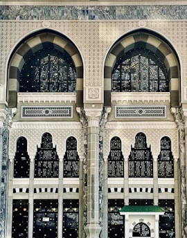 An intricate and ornate architectural facade featuring large arched windows with decorative patterns. The structure is adorned with geometric and floral motifs, displaying detailed craftsmanship in stone and glass. The design combines traditional and artistic elements, emphasized by columns and arches, creating a sense of grandeur.
