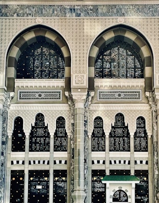 An intricate and ornate architectural facade featuring large arched windows with decorative patterns. The structure is adorned with geometric and floral motifs, displaying detailed craftsmanship in stone and glass. The design combines traditional and artistic elements, emphasized by columns and arches, creating a sense of grandeur.