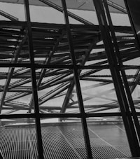 The image features a complex geometric structure composed of crisscrossing metal beams and a perforated metal facade. The design elements create a sense of depth and intricate detail, with light partially filtering through the spaces in between.