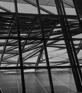 The image features a complex geometric structure composed of crisscrossing metal beams and a perforated metal facade. The design elements create a sense of depth and intricate detail, with light partially filtering through the spaces in between.