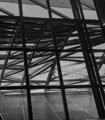 The image features a complex geometric structure composed of crisscrossing metal beams and a perforated metal facade. The design elements create a sense of depth and intricate detail, with light partially filtering through the spaces in between.