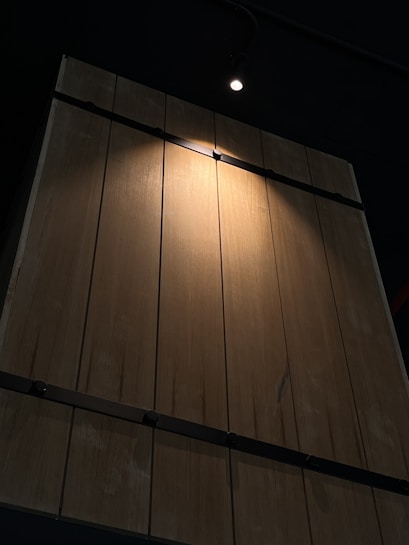 A wooden panel wall is illuminated by a single spotlight from above. The wooden planks are vertical and appear to be slightly weathered with natural wood grain visible. The spotlight creates a focused, bright area on the wooden surface while the surrounding area remains in shadows.