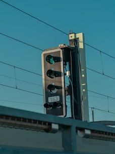 A close-up view of a railway signal light with a green light illuminated, set against a clear blue sky. Electric wires are visible in the background, indicating it's beside a railway track.