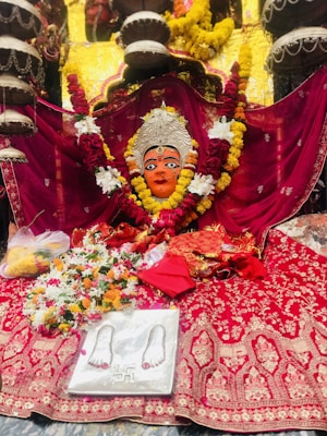 An ornate setup featuring a vibrant and detailed deity idol adorned with an intricately designed silver crown and surrounded by colorful flower garlands. The idol rests on a rich, embroidered red fabric. Additional elements include floral arrangements and a symbolic marking featuring feet impressions on a silver plaque.