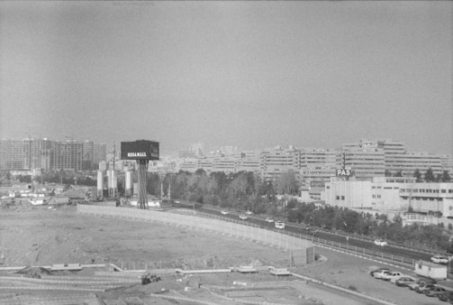 A black and white cityscape featuring a view of multiple mid-rise apartment buildings in the background and a large billboard with 'MEGA MALL' in the foreground. The scene includes a highway with several cars, some trees lining the road, and an open construction area.