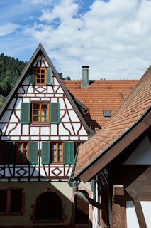 A historic half-timbered house with green shutters and intricate woodwork stands against a backdrop of lush forested hills. The steep, red-tiled roof and chimneys add to its traditional architectural style.
