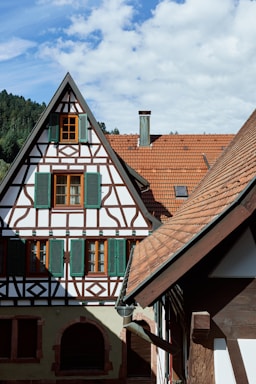A historic half-timbered house with green shutters and intricate woodwork stands against a backdrop of lush forested hills. The steep, red-tiled roof and chimneys add to its traditional architectural style.