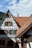A historic half-timbered house with green shutters and intricate woodwork stands against a backdrop of lush forested hills. The steep, red-tiled roof and chimneys add to its traditional architectural style.