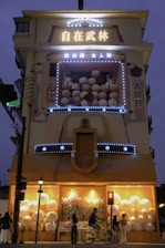 A building facade featuring illuminated signage with large Chinese characters and the words 'Women Street' in English. The structure includes a playful, oversized arcade game display filled with bouncy balls and two astronaut figures. The lower section showcases large spherical decorations and lights, while people walk and cycle by.