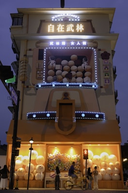 A building facade featuring illuminated signage with large Chinese characters and the words 'Women Street' in English. The structure includes a playful, oversized arcade game display filled with bouncy balls and two astronaut figures. The lower section showcases large spherical decorations and lights, while people walk and cycle by.