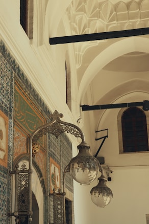 The image features an intricately decorated interior with ornate calligraphy and floral designs on a tiled wall. Large, antique-style hanging lanterns made of metal and glass are prominently displayed. The architecture showcases arched ceilings with geometric patterns, contributing to a historical and cultural atmosphere.