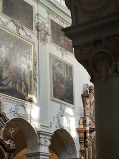 An ornate interior with intricate architectural details featuring columns and arches. Large oil paintings with gilded frames hang on the walls. Decorative moldings and sculptures adorn the space, and soft natural light filters through, casting gentle shadows.