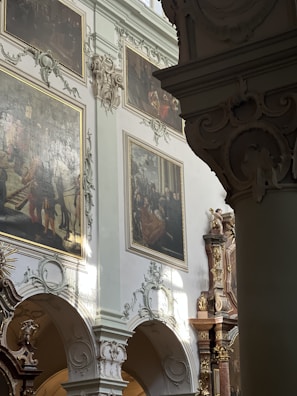 An ornate interior with intricate architectural details featuring columns and arches. Large oil paintings with gilded frames hang on the walls. Decorative moldings and sculptures adorn the space, and soft natural light filters through, casting gentle shadows.