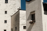 A rustic building with a white stucco exterior, featuring several small windows. A window box with vibrant red and pink flowers adds a touch of color against the plain facade. Shadows cast by the sunlight emphasize the texture of the walls, creating a tranquil yet slightly aged appearance.