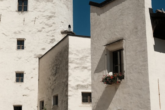A rustic building with a white stucco exterior, featuring several small windows. A window box with vibrant red and pink flowers adds a touch of color against the plain facade. Shadows cast by the sunlight emphasize the texture of the walls, creating a tranquil yet slightly aged appearance.