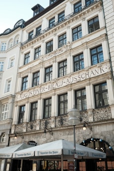 The image depicts an ornate, multi-story building with classical architectural elements. The facade features numerous windows and decorative ironwork. Prominently displayed is the text 'Augustiner-Bräu-Ausschank,' suggesting a beer hall or brewery. Canopies with matching branding are visible at street level, and a street lamp and outdoor seating are also present.