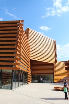 A modern architectural structure featuring horizontal wooden slats forming geometric patterns. The building's facade is made of glass and wood, creating an open and airy feel. There are a few people standing near the entrance, and the clear blue sky enhances the structure's warm, natural tones.