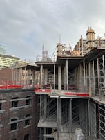 A partially constructed building with visible metal scaffolding and concrete pillars. The structure is surrounded by exposed brick walls and open spaces where windows might be installed. The sky is overcast, casting a neutral light over the scene.