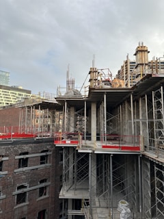 A partially constructed building with visible metal scaffolding and concrete pillars. The structure is surrounded by exposed brick walls and open spaces where windows might be installed. The sky is overcast, casting a neutral light over the scene.