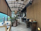A cozy outdoor cafe with wooden furniture and a bamboo awning creates a serene ambiance. Potted plants add greenery, while tables are set with napkins and candles. A small, organized bar is visible in the background.